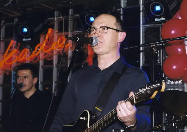 Wokalista z gitarą śpiewa na scenie, a w tle neonowy napis. Czas na koncert, a kiedy przyjdzie na ciebie czas tekst.