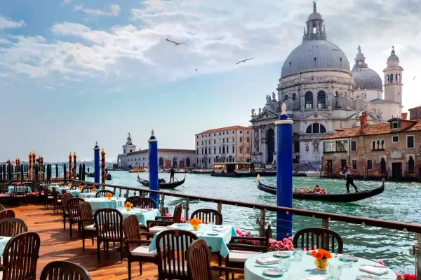 Tavoli apparecchiati su una terrazza con vista sul Canal Grande, gondole e la Basilica di Santa Maria della Salute. Un'ambientazione da sogno per i migliori ristoranti di Venezia.