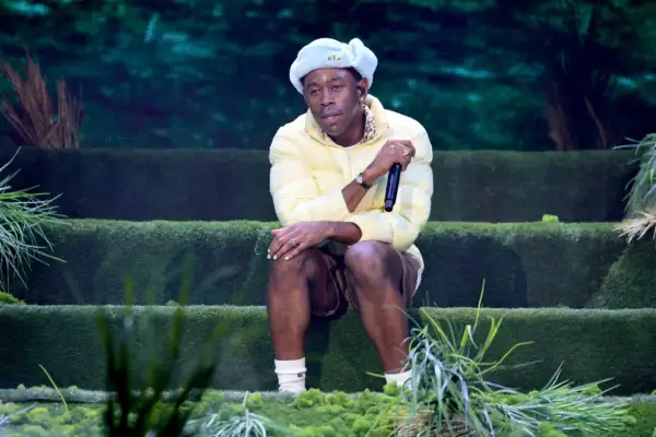 Igor, czyli Tyler, The Creator, siedzi na scenie w żółtej kurtce i jasnoniebieskiej czapce, trzymając mikrofon.