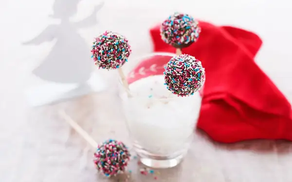 Färgglada cake pops med strössel står i ett glas med mjölk. En röd duk och en ängel i bakgrunden skapar en festlig stämning.