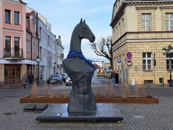 Pomnik z głową konia i niebieską szarfą, przypominający herb. W tle kamienice i ulica.