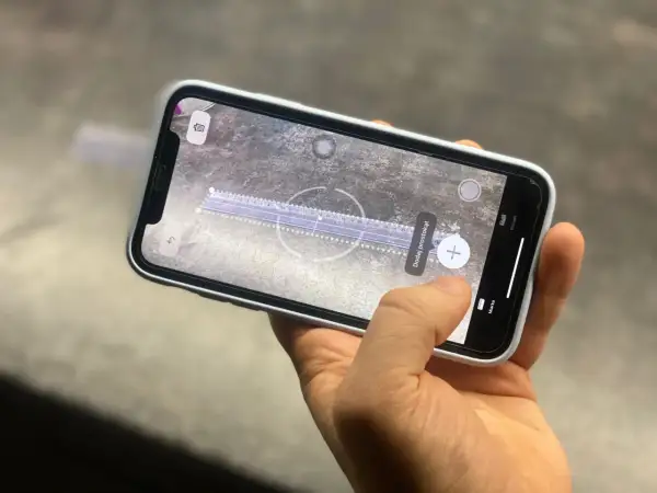 Dłoń trzyma smartfon, na ekranie którego działa aplikacja miarka. Wirtualna linijka mierzy odległość na powierzchni.