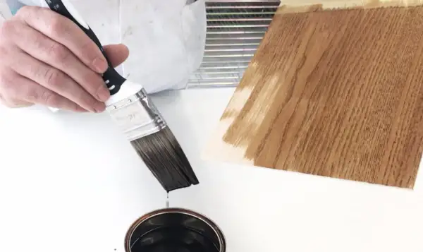 Pennello intinto in una latta di vernice scura, pronto per essere applicato su un campione di legno. Dopo l'impregnante, si mette la finitura.