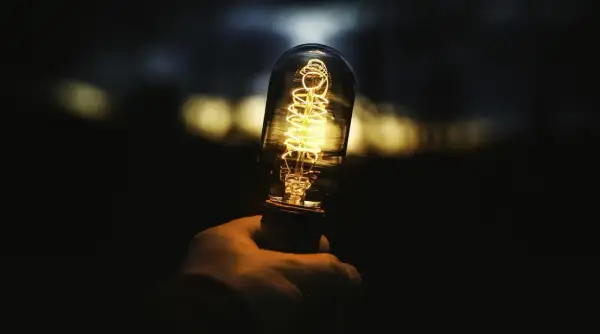 Dłoń trzyma żarówkę 1500 lumenów, której żarnik świeci ciepłym, żółtym światłem. Tło jest ciemne, z rozmytymi, jasnymi plamami.