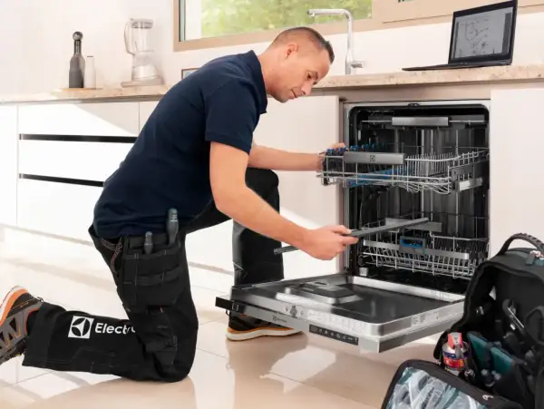 Serwisant w ubraniu z logo Electrolux naprawia zmywarkę. Electrolux to znana firma produkująca sprzęt AGD.