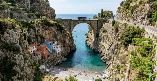 Un ponte ad arco domina un fiordo con case colorate aggrappate alla roccia. Il mare turchese invita a un bagno, mentre la strada panoramica, teatro del furore costiera amalfitana, accoglie bus e moto.