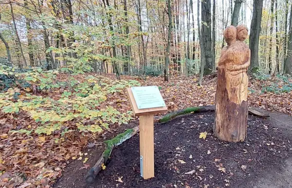 Holzschnitzerei zweier sich umarmender Figuren neben einem Schild mit der Aufschrift "Fatima Kapelle" im Herbstwald.