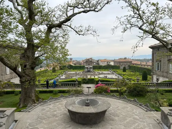 Giardino all'italiana con fontane e siepi geometriche, con un grande albero in primo piano. La serpara domina il paesaggio.