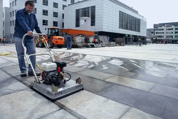 Uomo pulisce autobloccanti con idropulitrice in una piazza. L'acqua riflette la luce sui blocchi di pietra.