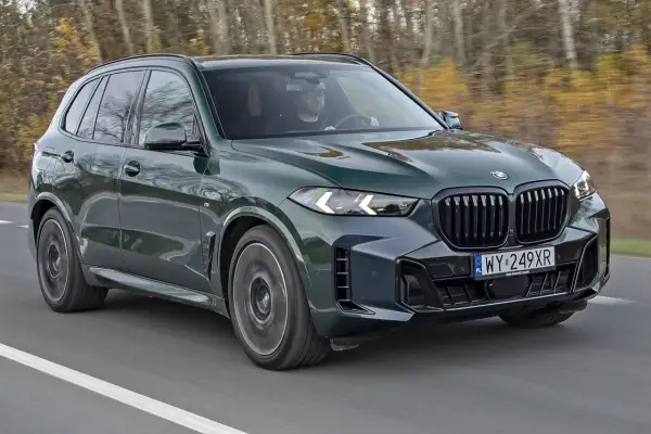 Nowe BMW X5 w ciemnozielonym kolorze mknie po drodze, otoczone jesiennym lasem. Kierowca w okularach pewnie trzyma kierownicę.