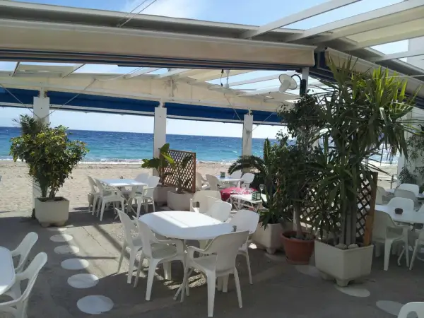 Terraza de restaurante en la playa. Mesas y sillas blancas bajo un toldo, con el mar azul de fondo. Un lugar perfecto para comer con agua amarga.