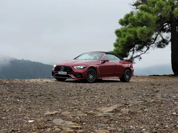 Czerwony kabriolet Mercedes-AMG CLE 53 na kamienistym podłożu, z drzewem i mglistymi górami w tle.