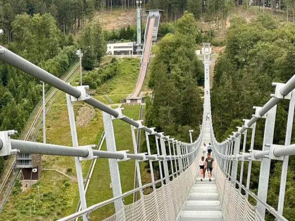 Menschen spazieren über den Willingen Skywalk, eine Hängebrücke mit Blick auf eine Skisprungschanze und bewaldete Hänge.