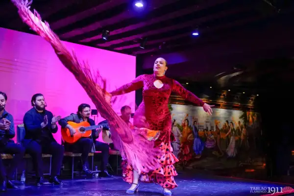 Bailarina de rumba flamenco con vestido rojo y volantes, moviendo un chal rosa. Músicos tocan guitarra y palmean al fondo.