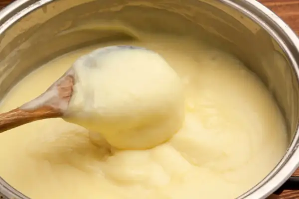 Une cuillère en bois prélève une généreuse portion de crème pâtissière onctueuse dans une casserole.
