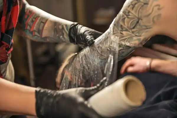 Manos enguantadas cubren un tatuaje recién hecho en el brazo con plástico. Aprende como cuidar un tatuaje recien hecho para una curación óptima.
