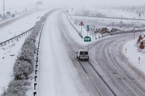 Nieve en la sierra de Madrid hoy. Una furgoneta blanca circula por una carretera nevada, con señales de tráfico y vegetación cubierta de blanco.