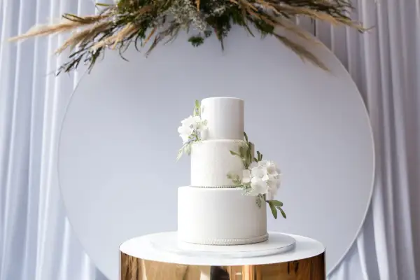 Eleganckie torty weselne wzory: biały, trzypiętrowy tort z delikatnymi kwiatami i koronkowym zdobieniem, prezentowany na złotym postumencie.