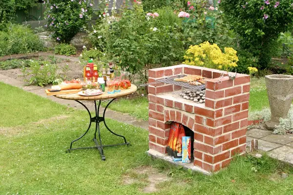 Ein selbstgebauter Ziegelgrill mit Würstchen und Kartoffeln, bereit für ein Gartenfest. Ein Tisch mit Salaten und Getränken steht daneben.