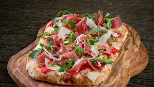 En avlång pinsa pizza med prosciutto, ruccola och parmesan på en träbricka.
