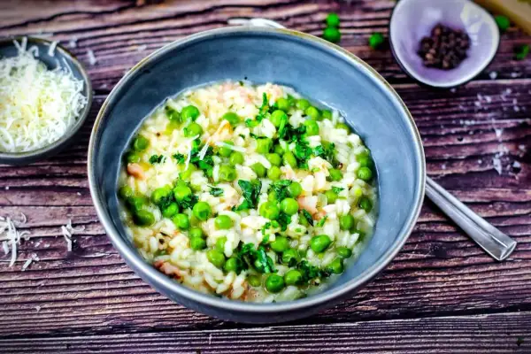 Ein cremiges Risi Bisi Rezept original, garniert mit frischer Petersilie und Erbsen. Daneben eine Schale geriebener Parmesan.