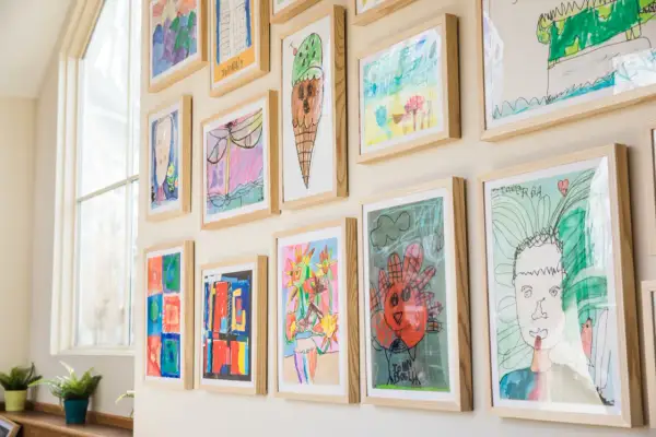 A vibrant kids art gallery wall showcases colorful drawings of ice cream, flowers, and portraits, each framed in light wood.