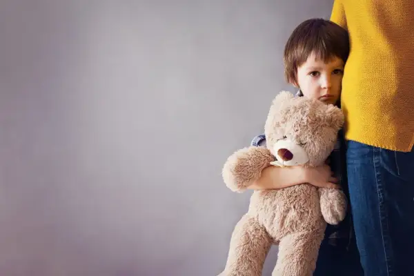 Chłopiec z pluszowym misiem przytula się do nogi dorosłego, wyrażając lęk separacyjny.