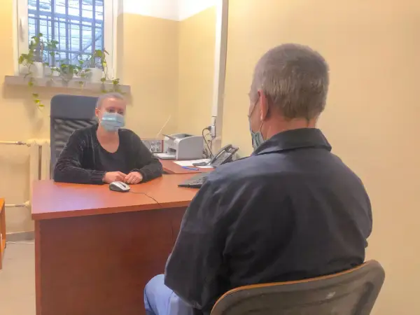 Psycholog więzienny w masce rozmawia z osadzonym. W tle okno z kratami i rośliny.