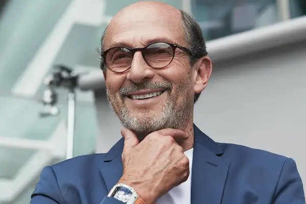 Uomo sorridente con occhiali, indossa un abito blu e un orologio Richard Mille, proprietario del brand.