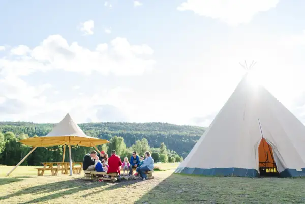 En familj njuter av en solig dag på alevi camping i Sverige. De sitter runt en lägereld under ett stort parasoll, med en tipi i bakgrunden.