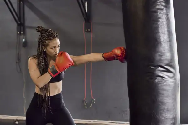 A woman with braided hair throws a punch at a heavy bag, proving that boxing tones arms and builds strength.