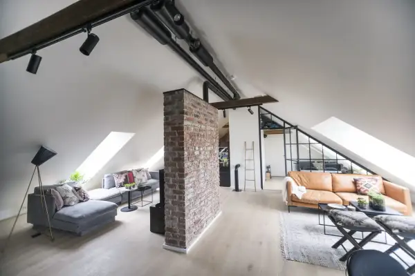 Un salon mansardé avec une cloison de séparation sous pente en briques apparentes, deux canapés, des luminaires industriels et des fenêtres de toit.