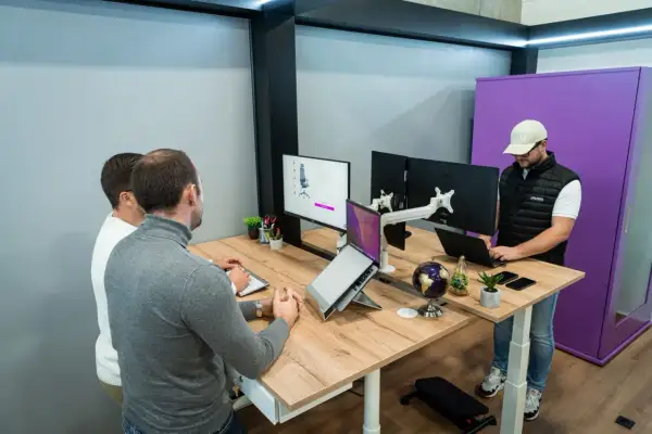 Trois personnes travaillent dans un bureau flex office moderne. L'un est debout devant un écran, les deux autres sont assis à un bureau.