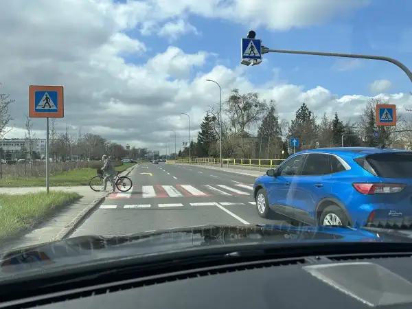 Niebezpieczne wyprzedzanie na pasach na drodze dwupasmowej. Niebieski SUV omija rowerzystkę stojącą przy przejściu.