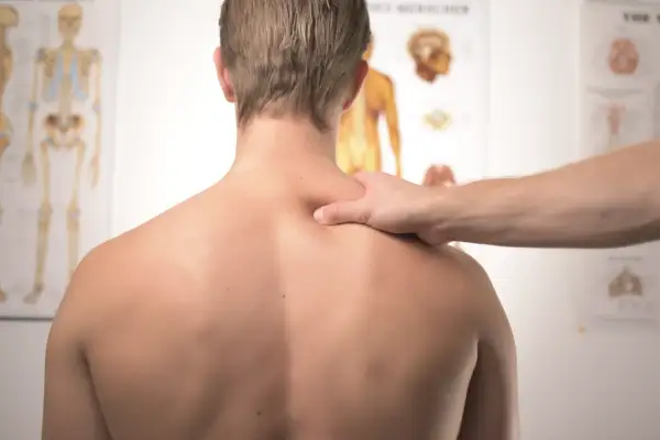 Dłoń terapeuty masuje bark mężczyzny na tle plakatów anatomicznych.