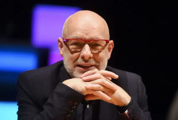 Brian Eno, eno musicista inglese, con occhiali rossi e mani intrecciate.