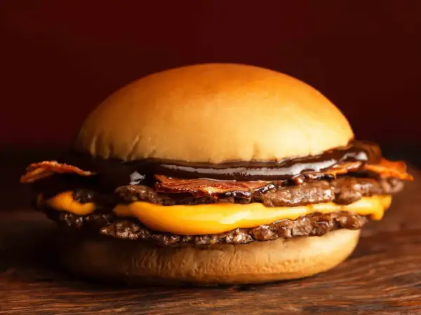 Gros plan sur un burger double viande, fromage fondant, bacon croustillant et sauce barbecue, sur une planche en bois.