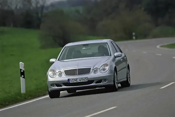 Srebrny Mercedes W211 z 2004 roku jedzie po krętej drodze wśród zielonych wzgórz.