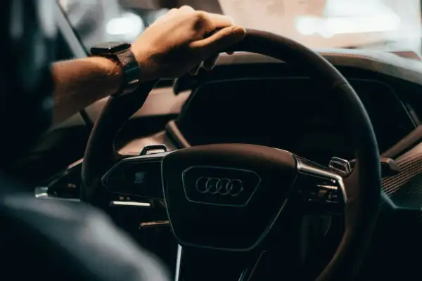 Dłoń na kierownicy Audi, z zegarkiem na nadgarstku. W tle widać fragment deski rozdzielczej, sugerujący wnętrze nowoczesnego samochodu, jak w tabeli z cenami rynkowymi samochodów używanych.