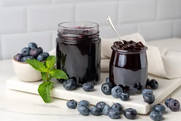 Due barattoli di marmellata di mirtilli, uno aperto con un cucchiaino, circondati da mirtilli freschi e foglie di menta.