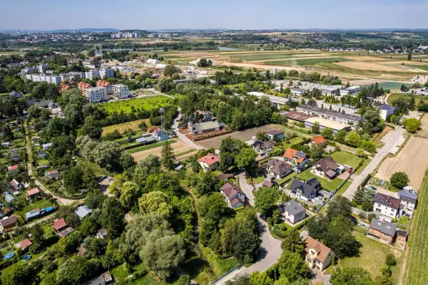 Osiedle domów jednorodzinnych na wzgórzach, otoczone zielenią i polami. W oddali widać zabudowania miejskie.