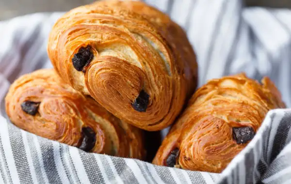 Tre deliziosi pain au chocolat dorati, con strati sfogliati e gocce di cioccolato fondente, adagiati su un canovaccio a righe.