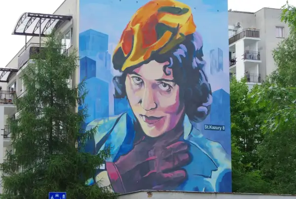 Mural przedstawiający Zofię Stryjeńską w żółtym beretce, z dłonią na piersi. Tło to błękitne, geometryczne budynki.