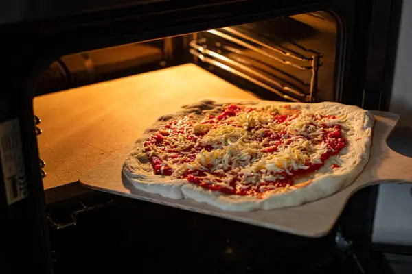 Pizza z sosem i serem na desce, gotowa do włożenia na rozgrzany kamień szamotowy w piekarniku.