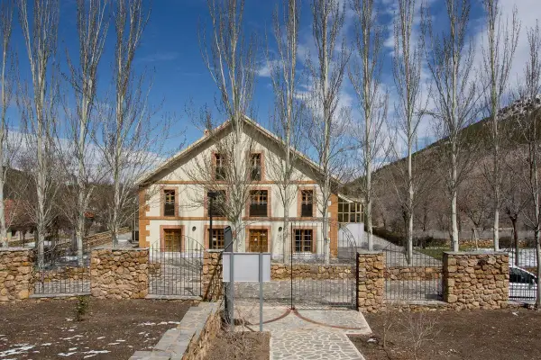 Edificio de piedra y madera con árboles secos delante. El camino de entrada está pavimentado con piedras. Las menas de la montaña se ven al fondo.