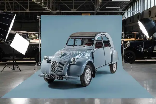 En ljusblå Citroën 2CV står i fokus, redo för fotografering. Bakom den syns andra klassiska bilar och studiobelysning.