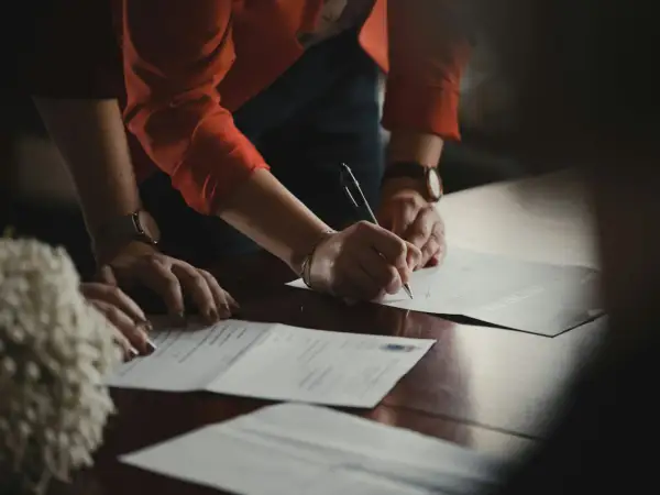 Dłonie podpisują dokumenty na drewnianym stole. Jedna z osób ma na nadgarstku zegarek, a druga nosi bransoletkę. W tle widać fragment pomponu. Klauzula RODO jest ważnym elementem umowy.