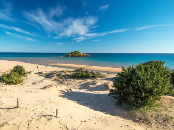 Dune sabbiose e mare turchese, un'isola rocciosa in lontananza. Un assaggio delle spiagge più belle Sardegna, con persone che passeggiano sulla riva.