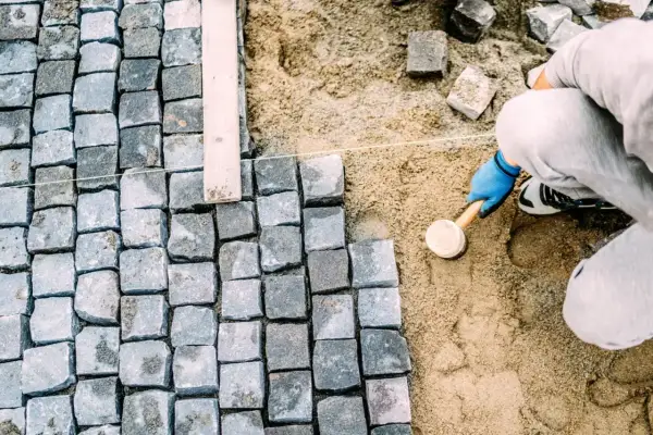 Pracownik w niebieskich rękawiczkach ubija kostkę brukową gumowym młotkiem, tworząc równą nawierzchnię.