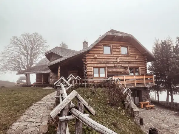 Drewniany dom w stylu góralskim, otoczony mgłą, zaprasza do odpoczynku w Beskidzie Wyspowym. Kamienna ścieżka prowadzi do przytulnego schroniska.